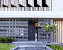 Modern building facade with horizontal screens and a touch of GB Breeze Blocks Linear Nickel featuring a central door, concrete and wood materials, a small flowering tree, and neatly trimmed grass in front.