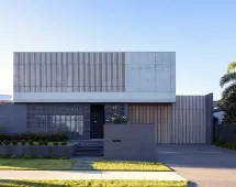 Modern two-story house with a minimalist design featuring GB Breeze Blocks Linear Nickel slatted panels, concrete walls, a flat roof, and manicured lawn. The exterior is finished in gray and beige tones.