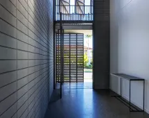 A modern hallway with tall GB Breeze Blocks Linear Nickel concrete walls, a polished floor, minimalist console table on the right, and a large slatted door that welcomes natural light and views of greenery outside.