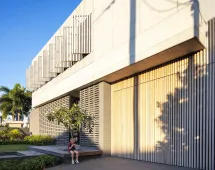 A modern building with vertical wooden and concrete slats casts shadows in sunlight. By the entrance, a person sits among lush greenery and palm trees, accented by GB Breeze Blocks Linear Nickel in the landscape.