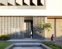 A modern facade featuring geometric brickwork, vertical wooden slats, a GB Breeze Blocks Linear nickel accent, a small tree, green lawn, and a child sitting on the sunlit steps near the entrance.