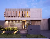 A modern two-story house with concrete and wood slats, geometric lines, large windows, and warm exterior lighting features GB Breeze Blocks Linear Nickel accenting the steps and landscaping at the front entrance at dusk.