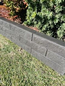A low Broughton Garden Edge Basalt retaining wall in dark gray separates a grassy lawn from a garden bed with green shrubs and red-tipped leaves. Bright sunlight highlights the wall and casts shadows on the grass.
