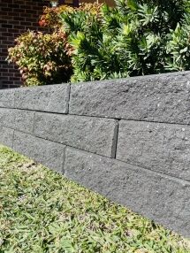 A close-up of a low, dark gray Broughton Garden Edge Basalt wall borders green grass with leafy shrubs behind it. In the background, there’s a brick building and part of a porch.