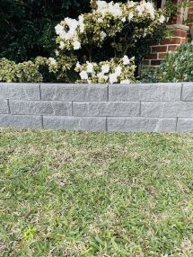 A low Broughton Garden Edge Basalt wall lines the lawn, with white flowering bushes behind it and a glimpse of red brick structure on the right.