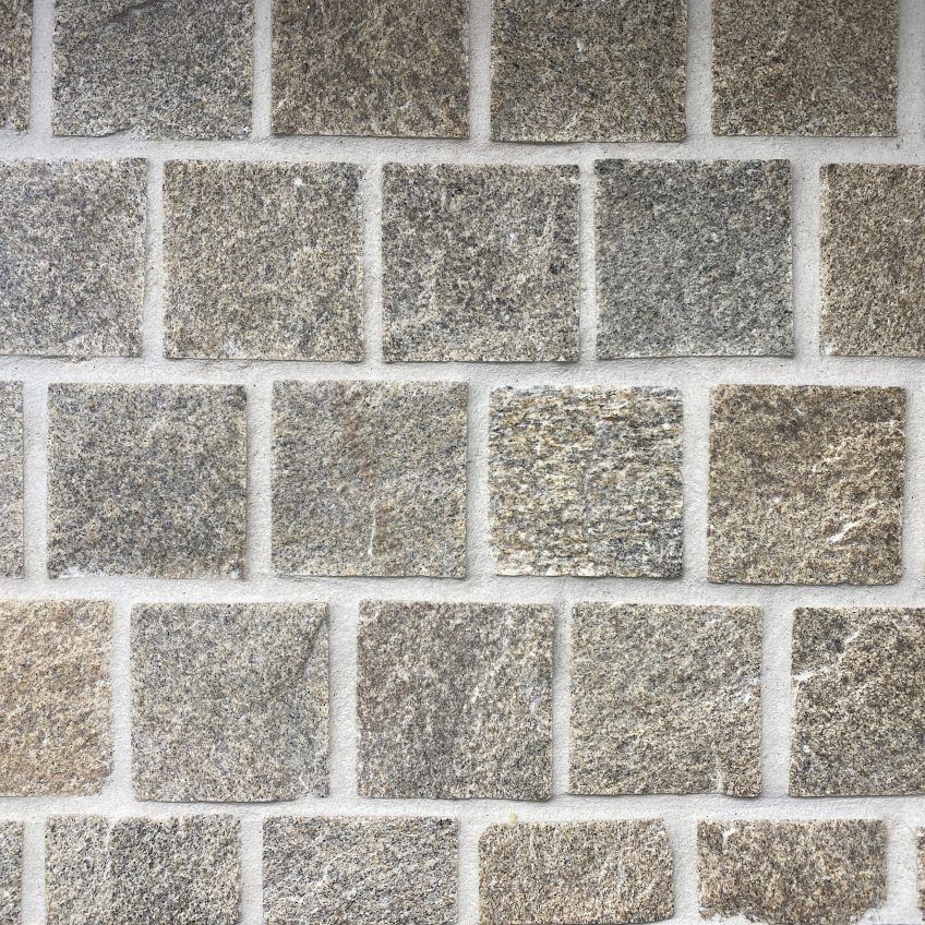 A close-up view of Tiger Skin Split Face Offset Cobbles reveals grey-brown bricks with a textured, natural stone surface arranged in a grid pattern, showcasing their rough finish and visible grout lines between each brick.