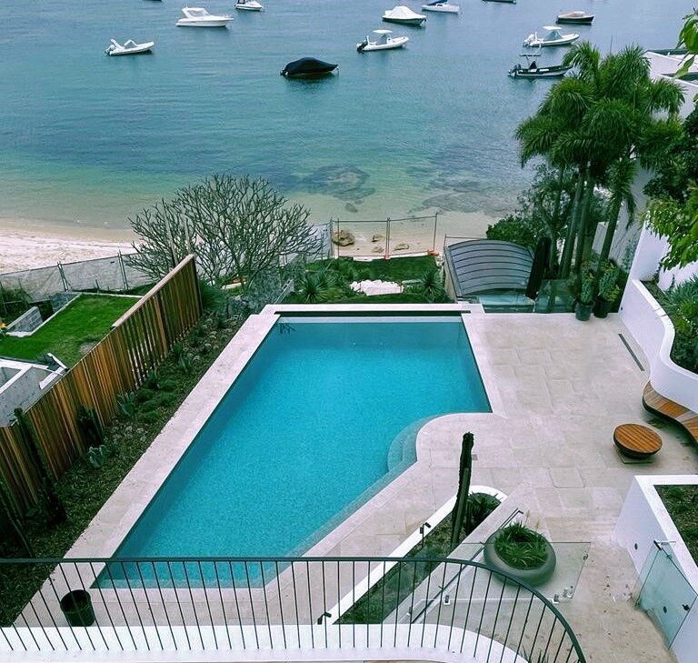 A modern, rectangular swimming pool adorns a tiled patio of Sinai Pearl S/blasted & Tumbled Paver, offering views of a tranquil bay with moored boats and a sandy shore. Lush greenery and palm trees frame the area, enhancing the peaceful waterfront ambiance.