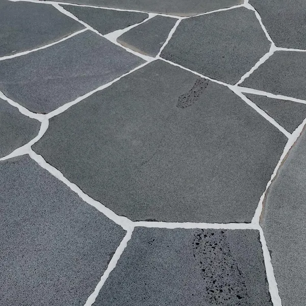 A close-up of a Crazy Paver made from Kashmira Bluestone showcases its irregular shapes forming a mosaic pattern. The dark grey stones contrast with the white mortar filling the gaps, and one stone bears a faint human footprint.