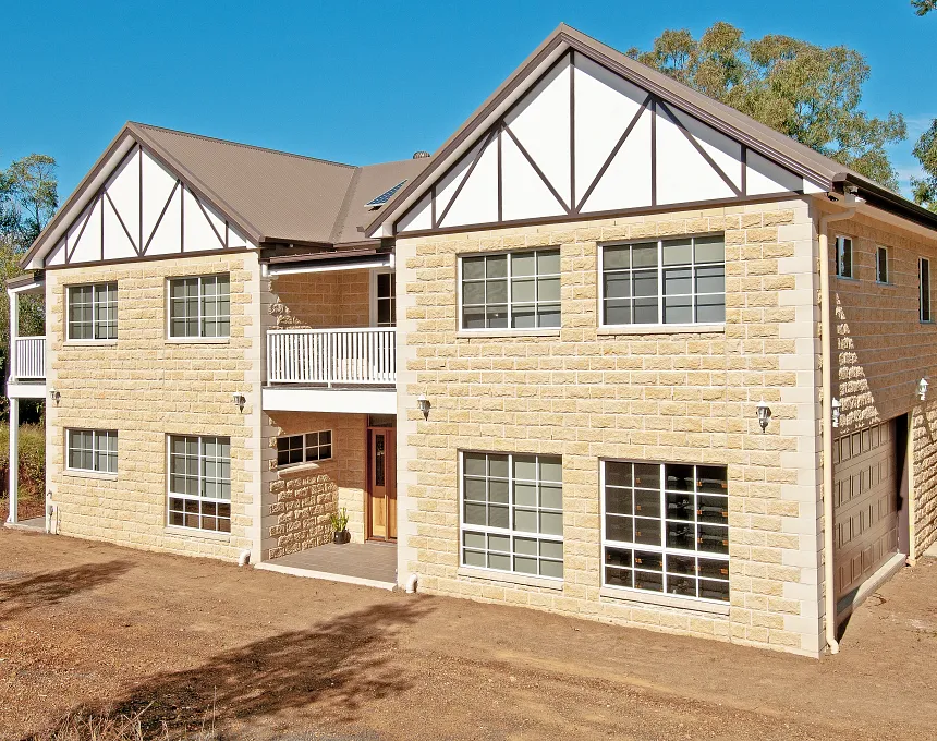 Two-story home features a GB Sandstone Rockface Sydney Blend exterior, dark trim, large windows, and a white balcony above the entrance. The sloped roof house sits on dirt with trees in the background.