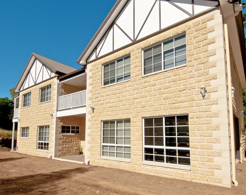 Two-story house with GB Sandstone Rockface Sydney Blend exterior, white trim, large windows, gabled roof, upper-level white balcony, and bare dirt around the home.
