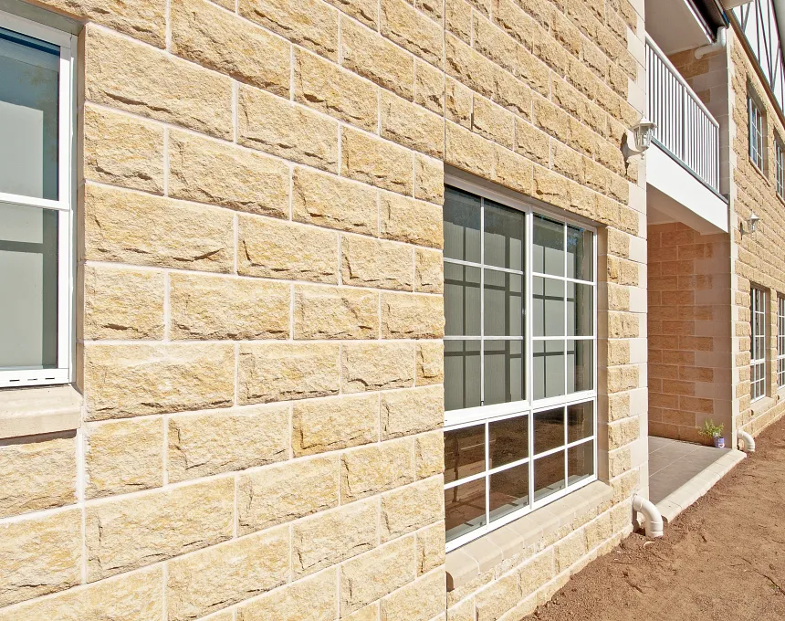 A close-up of a building exterior showcases large windows, a small balcony, and bare dirt beside the wall, all finished with GB Sandstone Rockface Sydney Blend bricks in light tan.