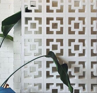 White decorative wall featuring the Breeze Block Double Square White design, showcasing a geometric cut-out pattern reminiscent of interlocking squares. Large green plant leaves stretch into the frame, offering a natural contrast to the modern aesthetic. A white brick wall is partially visible on the left.