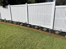 A neatly mulched garden bed with small, evenly spaced plants runs along a white slatted fence, bordered by a strip of rust-colored metal edging and surrounded by green grass.