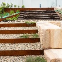 Modern landscaped garden with terraced steps made of large stones and rusted metal, bordered by gravel, small green plants, and a wooden staircase in the background.