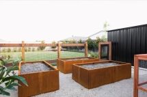 Three rectangular raised garden beds, bordered with FormBoss Metal Edging and filled with soil, are placed on gravel in a fenced backyard area next to a black shed, with a grassy lawn and houses visible in the background.
