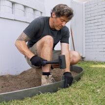 A man wearing gloves uses a rubber mallet to secure metal edging between a garden bed and grass in a backyard, kneeling on one knee.