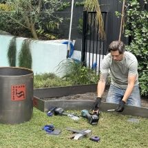 A man wearing gloves installs metal landscape edging in a garden. Tools and equipment are scattered on the grass nearby, and lush greenery surrounds the work area.