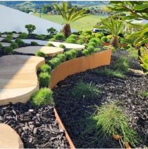 A landscaped garden with curved stone pathways, orange-brown edging, black mulch, clumps of green grass, small shrubs, and palm trees, set against rolling green hills in the background.