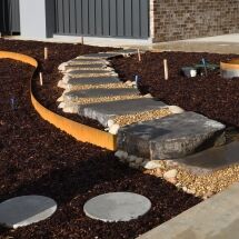 A modern garden path with large stone steps and a curved metal border, surrounded by brown mulch and small rocks, leading to a contemporary building. Circular concrete pavers are visible in the foreground.