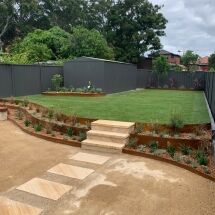 A backyard with a green lawn, small garden beds along the edges, and a grey shed at the back. Stone steps and a paved pathway lead up to the grass, with shrubs planted along the borders.