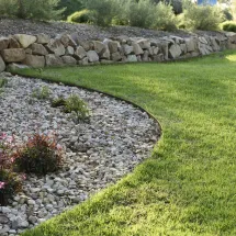 A landscaped garden with a curved border separates a gravel bed with plants from a neatly mowed green lawn. In the background, there is a low stone retaining wall and more greenery.