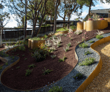 A terraced garden on a sloped hill features winding, curved retaining walls with various plants and shrubs, as well as mulched and gravel areas, shaded by tall trees with sunlight streaming through.
