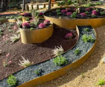 Curved, tiered garden beds with rust-colored metal edging display various small shrubs and flowering plants in different colored mulches: black, red, and gravel, in a landscaped outdoor area.