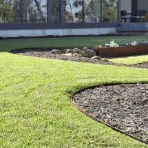 A modern landscaped garden features neatly edged green grass, a mulched garden bed with metal edging, and a rock feature near a contemporary building with large windows.