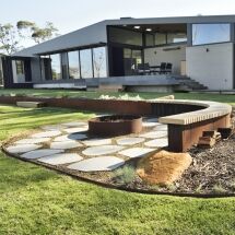 Modern house with large windows and a patio, featuring a landscaped yard with curved benches, a circular fire pit, stone pavers, green grass, and garden beds with rocks and plants.