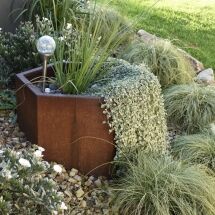 Hexagonal rust-colored planter with tall grass, silver trailing foliage spilling over the edge, a decorative glass orb stake, surrounded by gravel, green grasses, and white flowers in a lush garden setting.