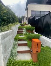 A modern outdoor staircase with concrete steps and FormBoss Metal Edging planters, surrounded by lush green grasses and ground cover, leads up to a contemporary house with black fencing and large windows under a cloudy sky.