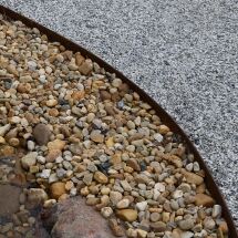Close-up view of a garden landscape with a curved metal edge separating tan river rocks from small gray gravel. The rocks and gravel create distinct textures and color contrasts.