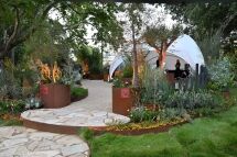A landscaped garden with stone and gravel paths, lush plants, metal planters, and a white dome tent in the background under large trees. A person stands inside the tent.