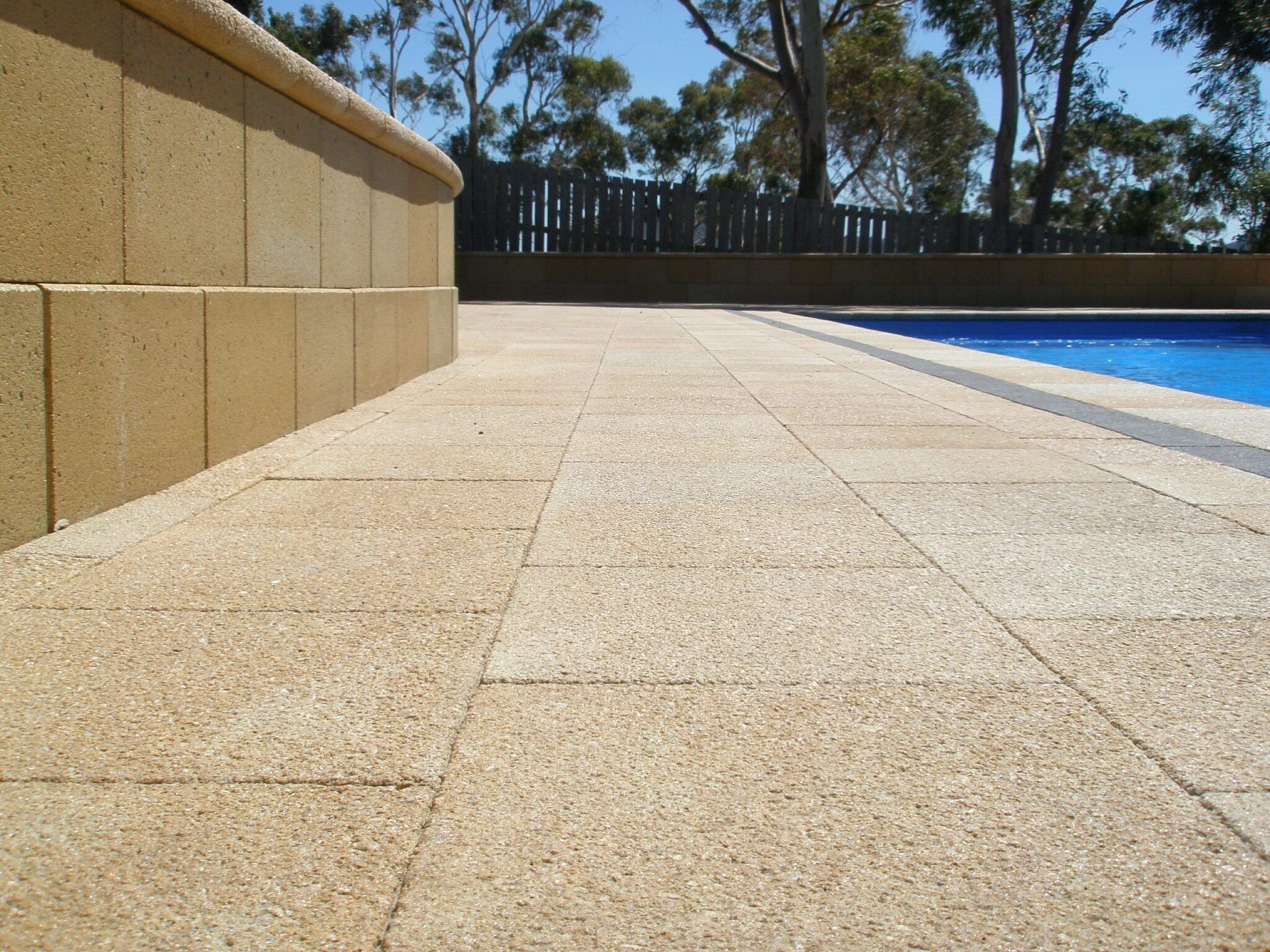 Close-up of a poolside area featuring Classic Stone Exposed Limestone tiles. A blue swimming pool is visible on the right, surrounded by trees under a clear blue sky.