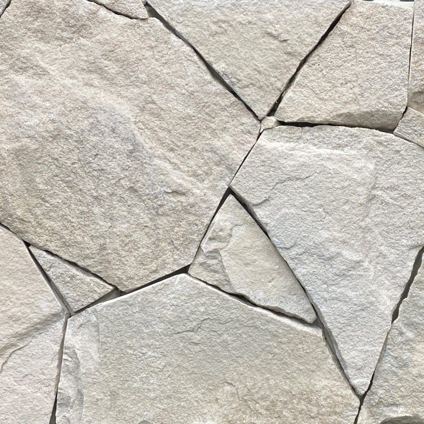 A close-up of the Avoca White Random Cladding Body highlights a textured stone wall composed of irregularly shaped, light grey stones that are seamlessly integrated. The rough surface exhibits natural variations and patterns throughout the stones.