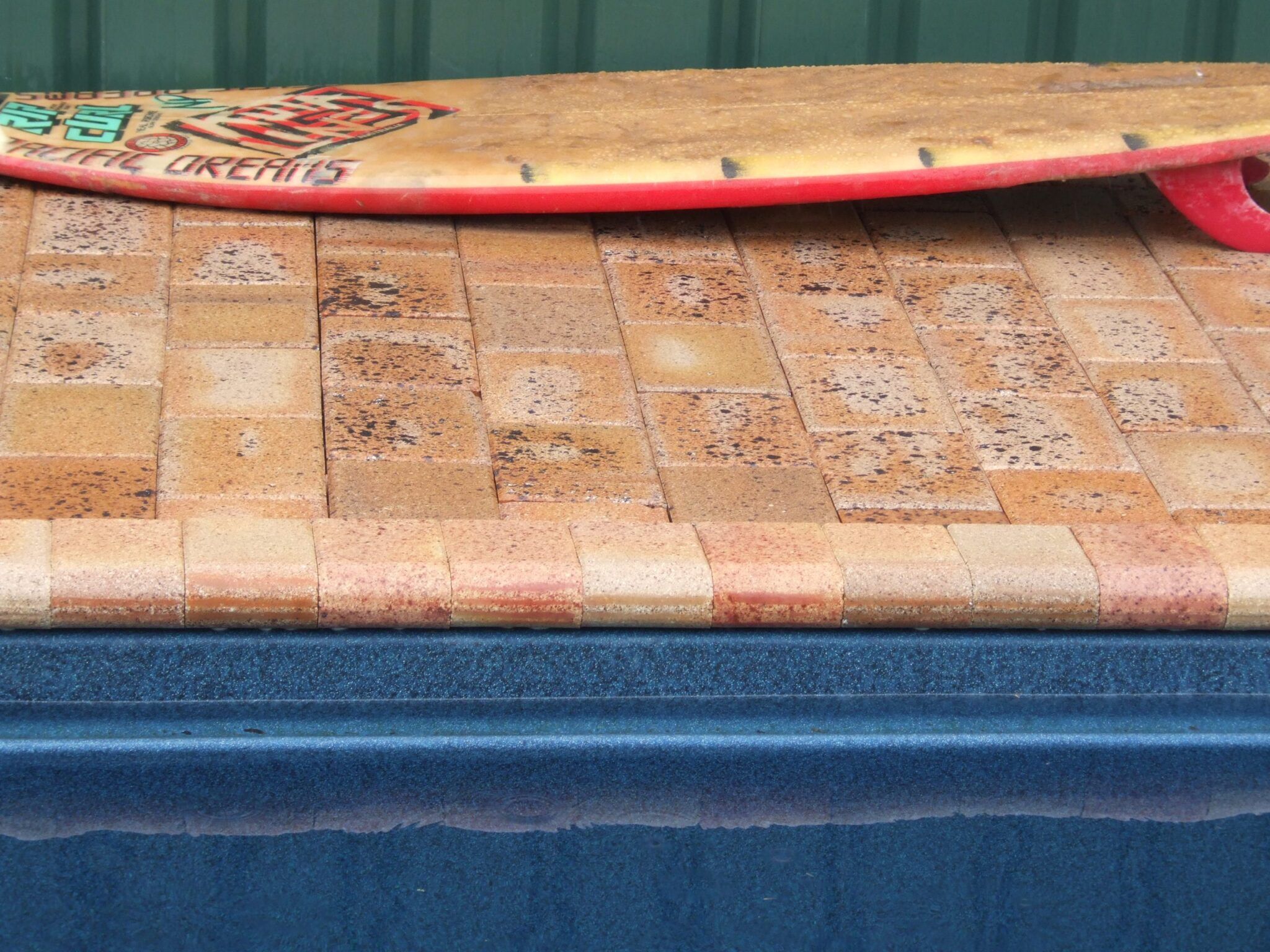 A surfboard with a colorful design is resting against a green wall on a surface tiled with Cobble Coper, near the edge of the blue swimming pool. The tiles are a blend of beige and brown tones.