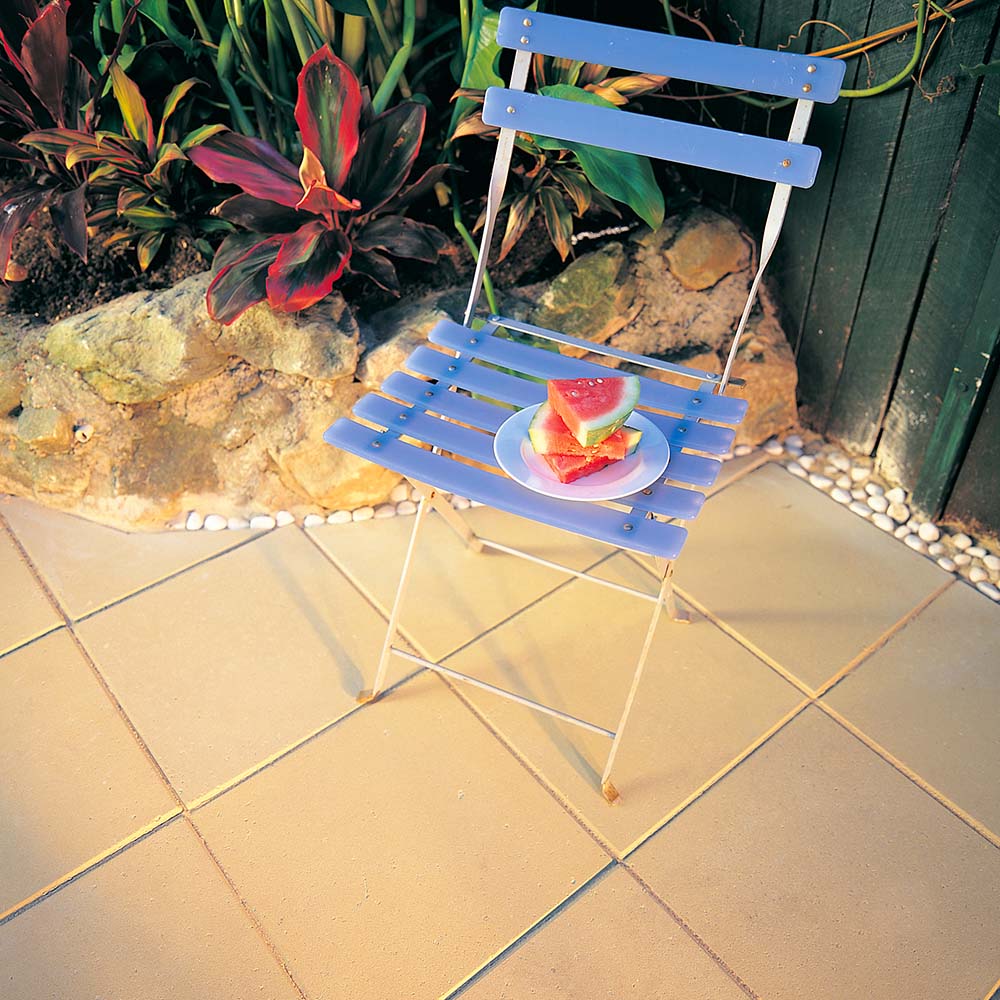 A blue wooden chair on the patio, near an Esplanade 400 Sand garden filled with green plants and rocks, holds a white plate with sliced watermelon. The light brown tiles perfectly complement the tranquil setting.